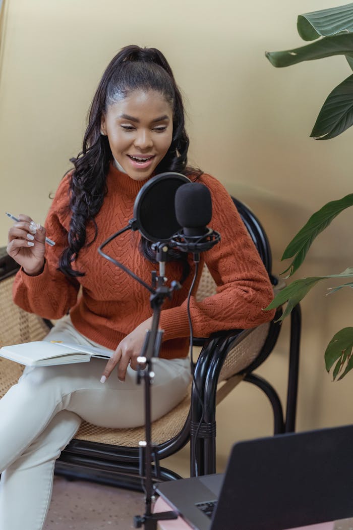 services-03 Positive African American female recording podcast with microphone while sitting in chair with notebook in light room with modern laptop